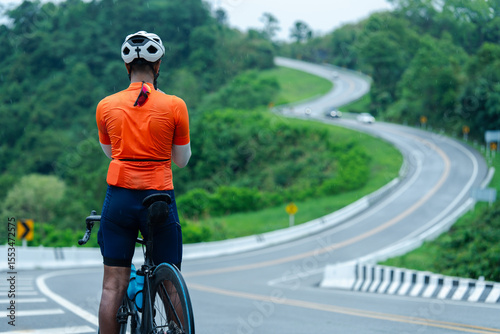 A cyclist in an orange jersey pauses on a winding road surrounded by lush greenery and trees.