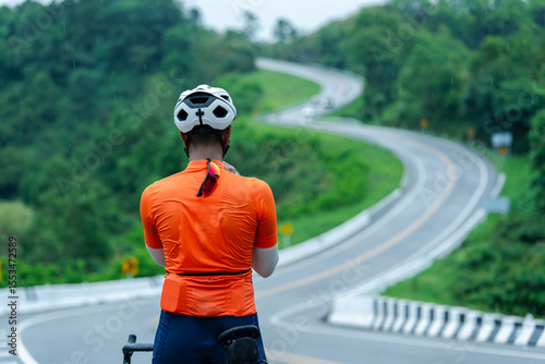 A cyclist in an orange jersey and helmet pauses on a winding road surrounded by lush green forest.