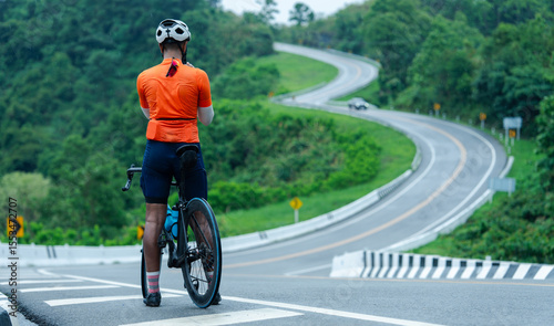 A cyclist in an orange jersey stands at the start of a winding road surrounded by lush greenery, ready to begin a challenging ride through a scenic landscape.