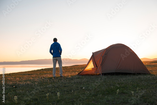 Man standing next to set up tent at a camping site by a highland freshwater lake surrounded by high mountains at sunset. Song Kul Lake, Kyrgyzstan, Central Asia. Road trip, travel camping concept.