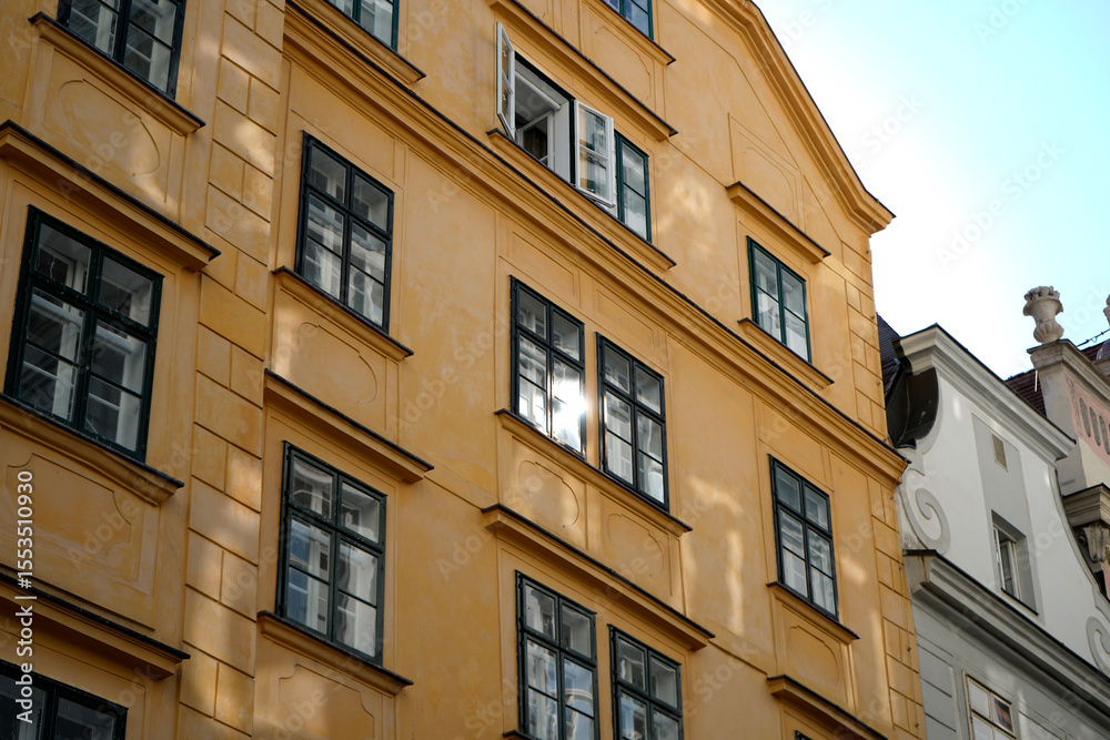 Naklejka premium Yellow urban facade with rhythmic windows and one open shutter. Glass reflects daylight softly. Elegant symmetry and warm color highlight traditional European style.