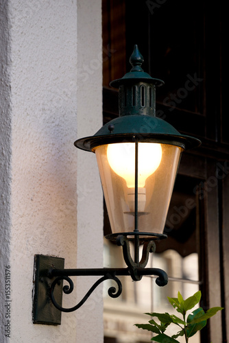Wall mounted lantern. Glowing vintage bulb. Iron street fixture. Textured white wall.

