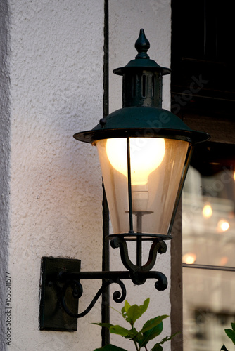 Wall mounted lantern. Glowing vintage bulb. Iron street fixture. Textured white wall.

