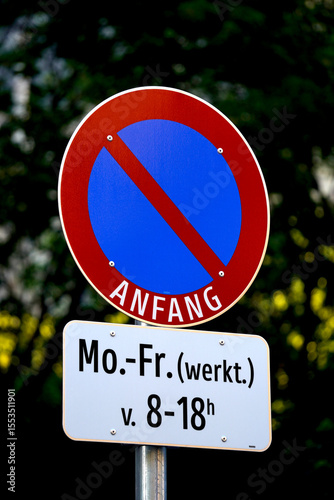 Close-up of Austrian no parking traffic sign with blue and red circle and time restriction. Clear daylight image with green tree background and sharp typography.

