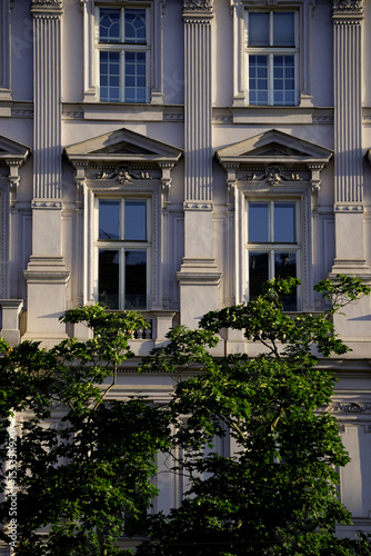 Classic baroque-style facade with tall columns, decorative window frames and sunlight through green tree. Elegant architecture and detailed ornamentation in historic context.

