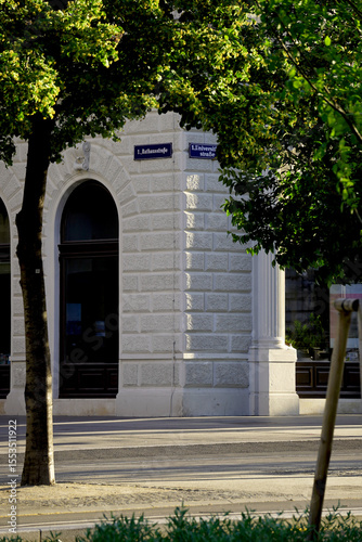 Historic European corner building with white stone facade and classic arched windows. Sunlight and tree shadows create calm urban scene at the street intersection.

