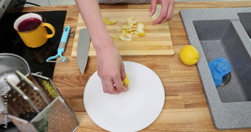 Female hands arrange lemon peel on white plate near fresh fruit and wooden board. Sharp knife rests nearby suggesting cooking in progress slow motion