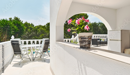 Bright Mediterranean-style terrace framed by white arches and balustrades. Relaxing outdoor setting with lush green trees in background during summer sunny day