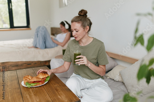Middle aged woman enjoying healthy lifestyle drinking green smoothie and eating fresh sandwich in cozy home environment focused on wellbeing and fitness