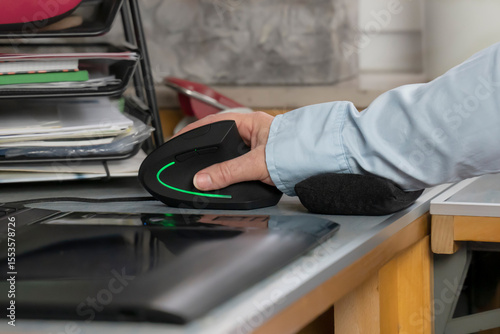 Ein Arbeitsplatz mit Computer. Auf dem Schreibtisch ist eine Hand in entlastender Position mit einer ergonomischen Maus. Neben der vertikalen Maus unterstützt ein kleines Kissen das Handgelenk.