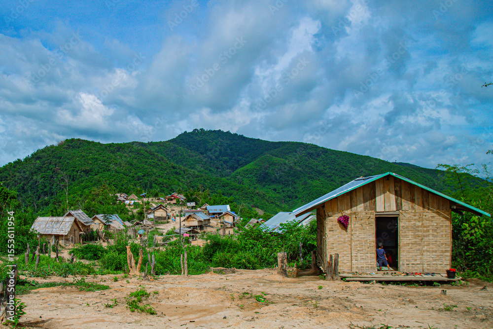 Fototapeta premium A traditional bamboo house with roof stands in the foreground, overlooking a sprawling rural village nestled within a valley of lush green mountains. 