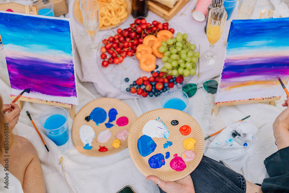 Fototapeta premium Close-up of artistic hands painting on canvases, vibrant palette includes purple, blue, red, and white. Picnic setup with champagne glasses, fresh fruit, and art supplies creates colorful creative mom