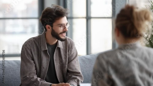 Young man in glasses having a serious conversation with a woman in a bright modern room, suggesting a therapy, counseling, or personal talk session.