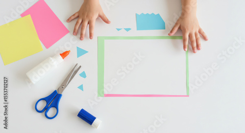 children's craft project with paper shapes and colorful materials. child's hands on white background surrounded by scissors, glue, and vibrant paper pieces for creative activity