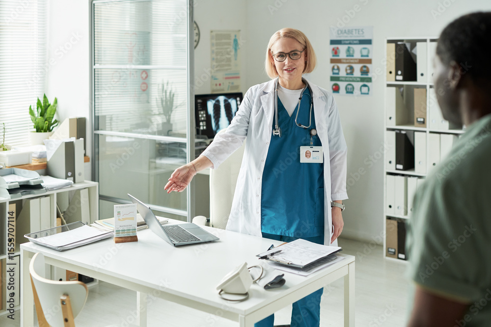 Obraz premium Middle aged Caucasian female doctor welcoming middle aged Black man patient into medical office, gesturing toward desk with medical documents and digital tablet visible on table