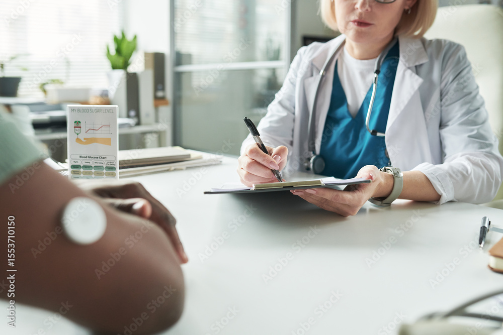 © pressmaster - Middle aged Caucasian female doctor consulting Black adult patient with diabetes, writing notes on clipboard while patient with glucose monitor on arm sitting across medical office desk
