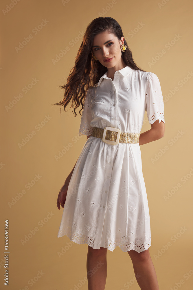 Fototapeta premium Chic young woman in a short-sleeve white eyelet dress with a woven belt and floral earrings, posing gracefully against a warm beige background. Perfect for fashion, beauty, and lifestyle themes.