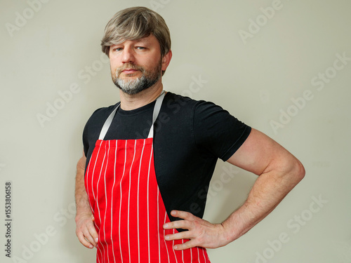 Portrait of professional butcher or fishmonger in black shirt and red and white classic traditional apron, light color background. Male model in his 40s with grey beard and hair. Hands on hip.