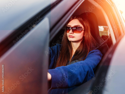 A young inexperienced woman is driving a car with her sunglasses on. She is wearing a blue hoodie. High cost of insurance for young drivers concept. Travel and tourism concept. Sun flare