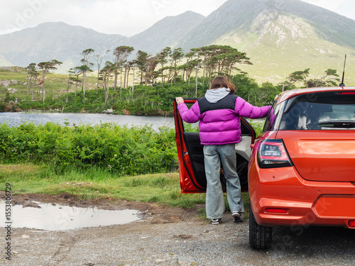 Young teenager girl standing by small red sport car and enjoys view of Twelve pines island and mountains in Connemara, county Galway, Ireland. Famous tourist area with amazing nature scenery.