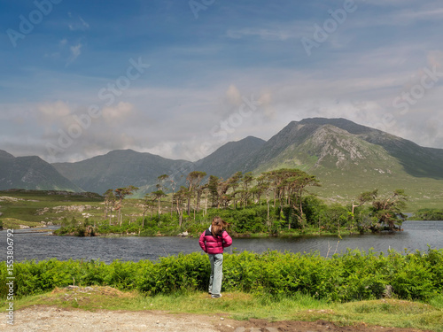 Young girl tourist looking at stunning nature scenery of Connemara, county Galway, Ireland. Lake, Twelve pines island and tall mountains and blue cloudy sky. Travel and sightseeing. Irish nature.