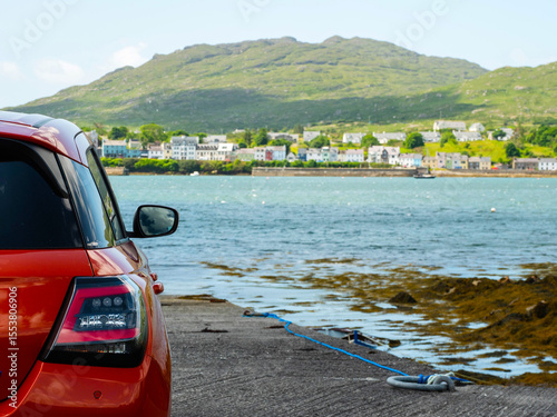 Small sport red car is parked by the ocean with stunning view on nature scenery and Roundstone town in the background with Errisbeg Mountain, county Galway, Ireland, Travel and tourism.
