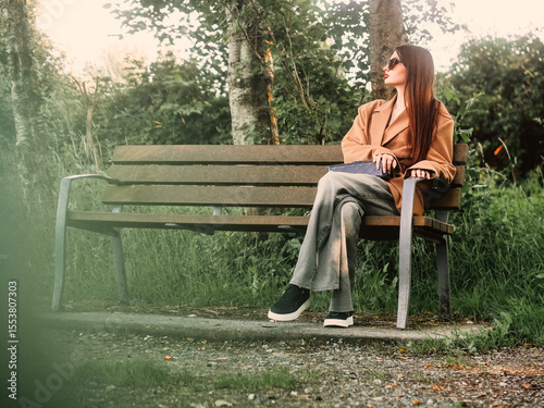 Young and stylish teenager girl sitting on a bench in a park alone. Waiting for date concept. The model wears trendy brown jacket and dark glasses and looks calm and elegant. Selective focus.