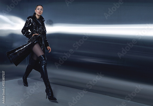 Fashion model in black leather trench coat and thigh-high boots posing in futuristic, metallic interior. Elegant editorial fashion photoshoot with dramatic lighting.