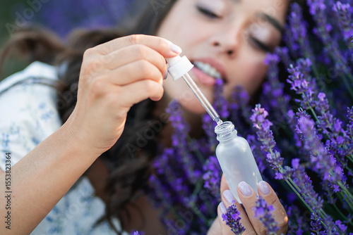 Closeup of a young beautiful dark haired woman applying serum to her face near lavender. Holding a cosmetic jar with a pipette. Essential oil.