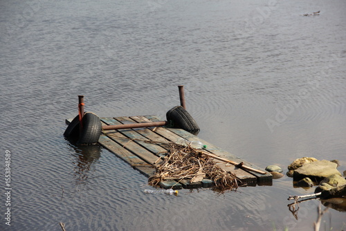 Partially Submerged Wooden Raft with Tires and Debris Floating in Calm Water Near Rocks and Shoreline