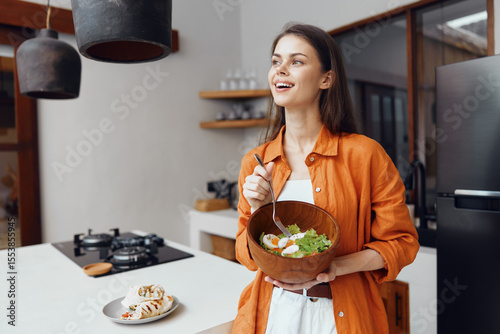 Healthy eating woman in an orange shirt joyfully holding a salad bowl in a modern kitchen setting, with fresh ingredients and natural light enhancing the atmosphere