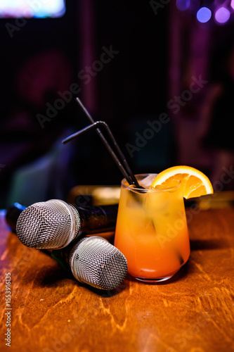 Orange cocktail with a straw on a bar counter next to two microphones. Cozy karaoke bar or party vibe