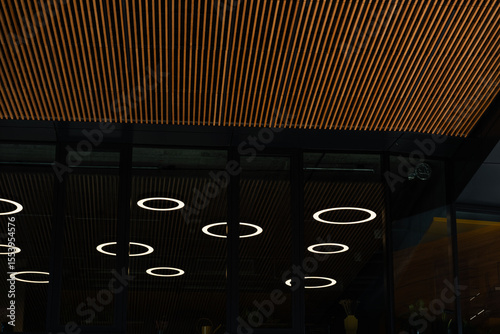 Photo shows a contemporary ceiling with wooden slats and circular LED lights reflected in glass. The image highlights modern architectural design and interior lighting solutions.
