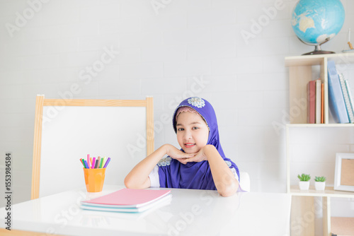 Children Muslim girl in purple hijab sitting at white desk with colorful markers and notebooks, smiling at camera in bright study room with whiteboard and bookshelves, Asian student in relax time.
