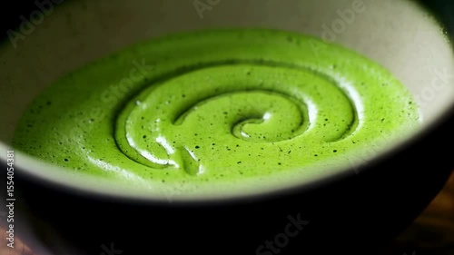 Close-up of vibrant green matcha tea being whisked in a bowl, creating swirling patterns, with a rustic wooden background
