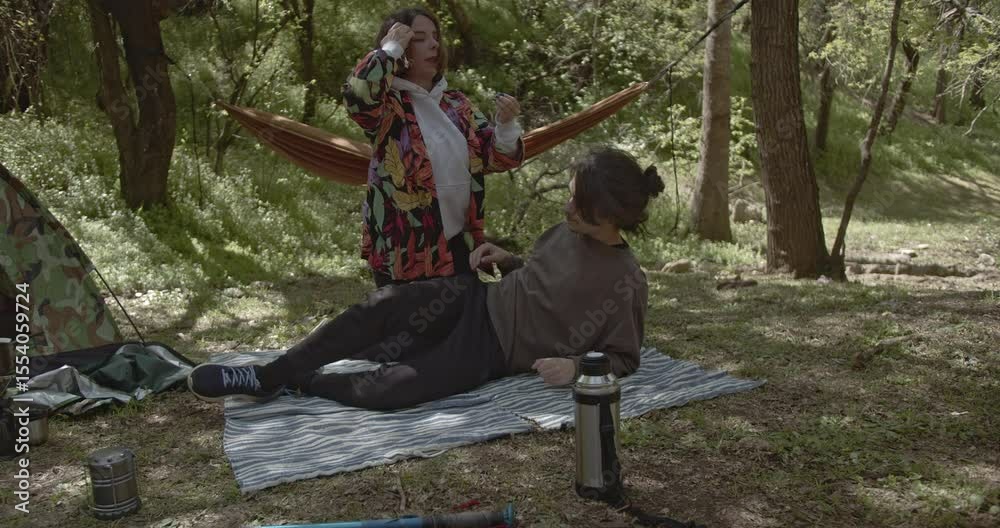 Friends relax and enjoy their camping trip in the woods. They stretch, fix their hair, and take in the serene atmosphere.