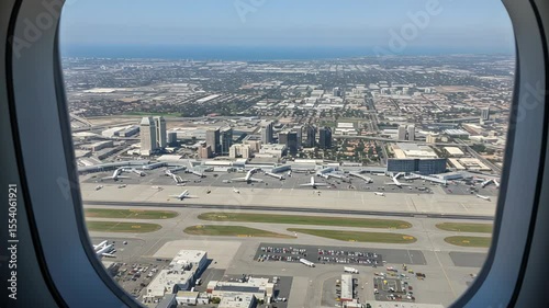 Aerial View From Airplane Window Showcasing Cityscape With Buildings, Roads and Parkings Full With Cars, An Airport Runway And Blue Sky