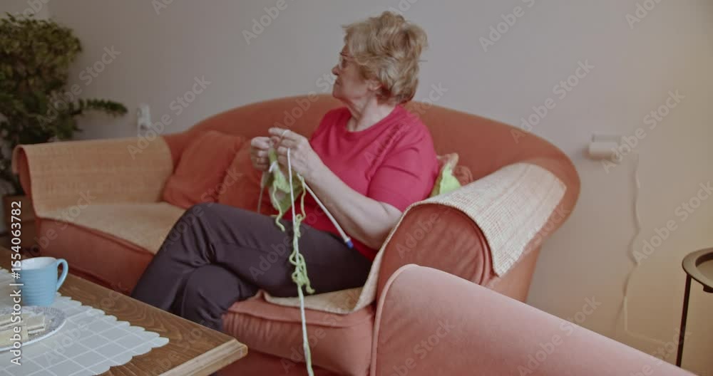 Elderly woman enjoys her retirement, knitting on the sofa with a cup of tea and biscuits nearby. She finds peace and comfort in this hobby.