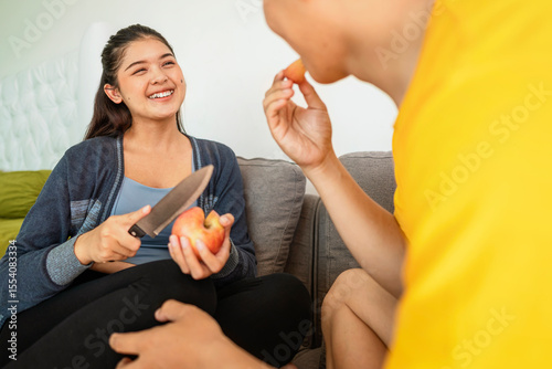 Indonesian southeast asian wife with a knife serves a slice of fruit to her husband while sitting on the sofa at home. Healthy eating concept