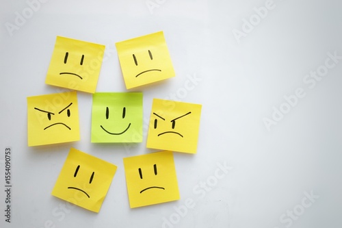 A green sticky note with a smiling face surrounded by yellow sticky notes with frowning and angry faces, symbolizing positivity among negativity