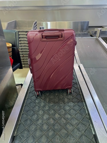 red Suitcase on Airport Baggage Claim Conveyor Belt. red hardshell suitcase stands prominently on a black rubber conveyor belt at an airport baggage claim.