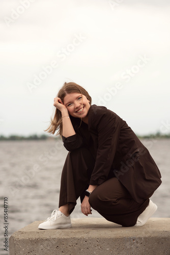 Smiling woman in brown suit leaning on railing near water. Natural feel with soft light. Casual, confident pose and expression add genuine and approachable vibe
