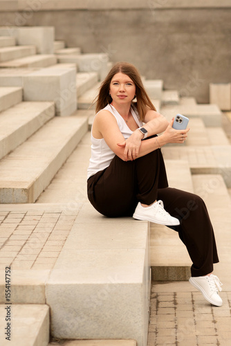 Young woman sitting on stairs holding phone and looking away outdoors. Natural feel with soft light. Casual, confident pose and expression add genuine and approachable vibe