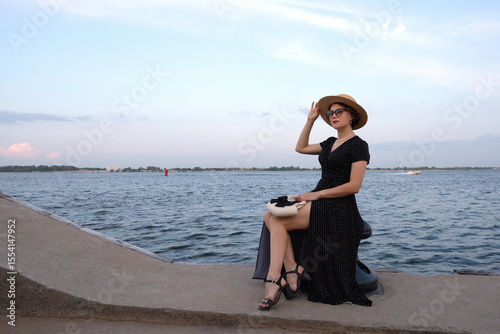 Woman in polka-dot dress poses by the waterfront promenade. Lady with beautiful long legs poses on the bank of a body of water. Summer vacation concept