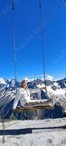 A woman in a white tracksuit is sitting on a swing in front of a mountain,looking into the distance.