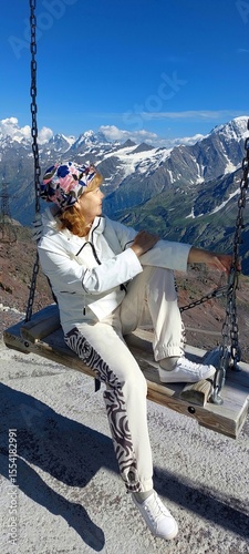 A woman in a white tracksuit is sitting on a swing in front of a background of snowy mountains.She is looking at the mountains.