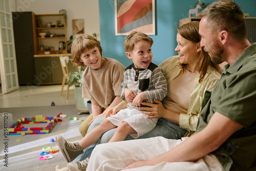 Caucasian middle aged woman and man sitting on sofa interacting with two Caucasian boys, one child and one teenager, smiling and laughing together in home setting with toys visible
