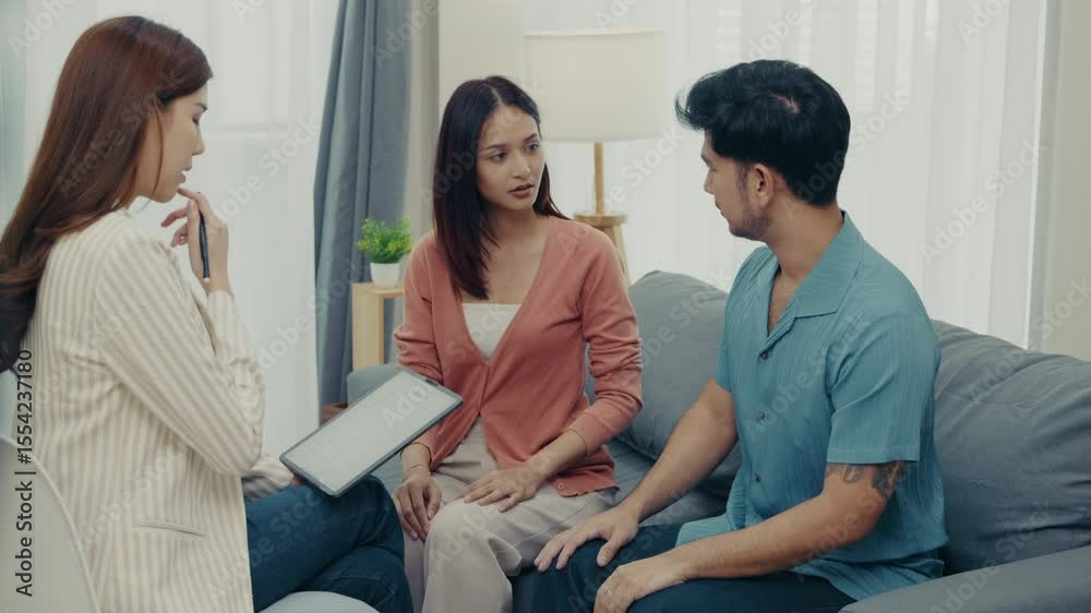 An Asian couple communicates openly during a therapy session at home with a female counselor. The man leads the conversation while the woman listens attentively in a calm atmosphere. An Asian couple communicates openly during a therapy session at home with a female counselor. The man leads the conversation while the woman listens attentively in a calm atmosphere.