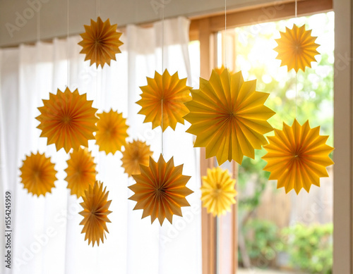 Numerous bright yellow paper starburst decorations of various sizes hang delicately by white strings in front of a large window with sheer white curtains, creating a cheerful, sunlit ambiance. photo