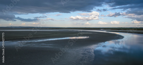 Calm waters reflect a dramatic sky at low tide near a tranquil estuary in the late afternoon
